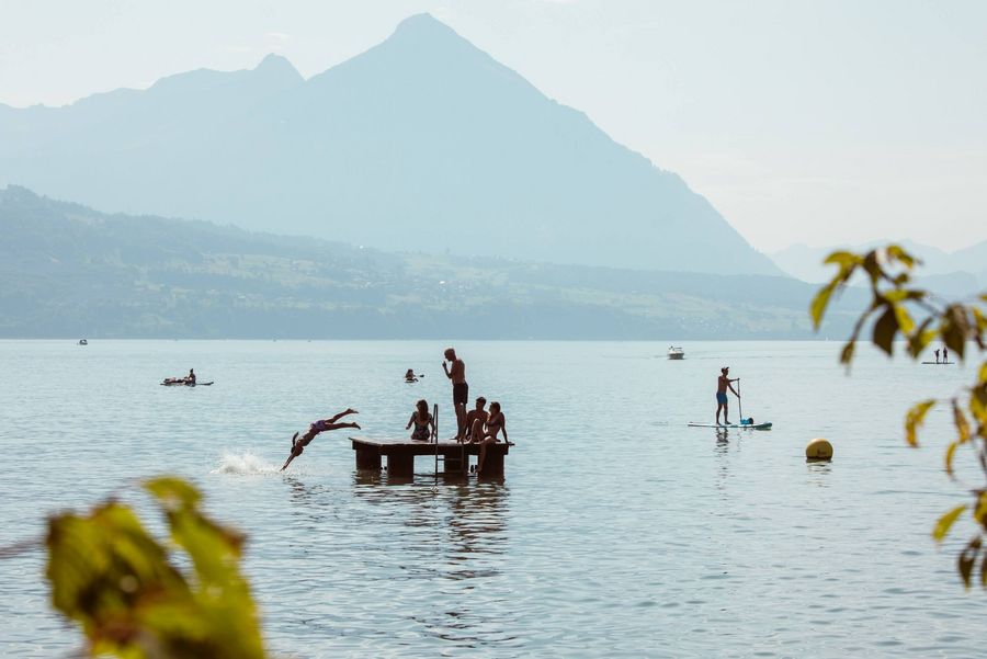Schülerbad Neuhaus mit Personen, die auf dem Floss stehen oder in den Thunersee springen sowie Stand-up-Paddler und Boote im Hintergrund